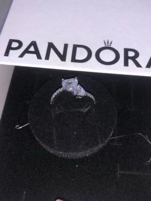 Silverring med hjärtformad sten Pandora - Säljer en elegant silverring från Pandora med en stor hjärtformad klar sten i mitten och mindre gnistrande stenar längs bandet. Ringen levereras i originalask och har en klassisk, romantisk design som verkligen sticker ut.