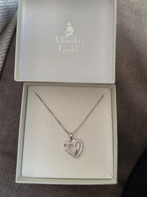 Halsband med hjärthänge i silver - Snyggt halsband från Albrekts Guld med ett hjärthänge i silverfärg. Hänget har en öppen hjärtform och är dekorerat med små gnistrande stenar längs kanten. Kedjan är tunn och elegant, perfekt för att lyfta vilken outfit som helst. Aldrig använt och det kommer i orginalförpackningen💓