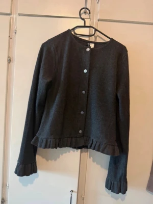 Mörkgrå kofta med volangdetaljer - Snygg mörkgrå kofta med rund halsringning och knappar framtill. Koftan har volangdetaljer längs nederkanten och vid ärmsluten som ger en feminin touch. Långärmad och i mjukt material, perfekt att slänga på sig över en topp.