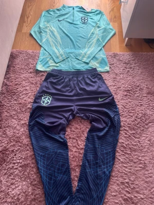 Nike Brasil träningsset långärmad - Snyggt träningsset från Nike med långärmad ljusblå tröja och mörkblå byxor. Tröjan har Brasil-logga och dragkedja vid halsen, medan byxorna har matchande logga och mönster i blått och grönt. Perfekt för fotbollsträning eller gymmet.