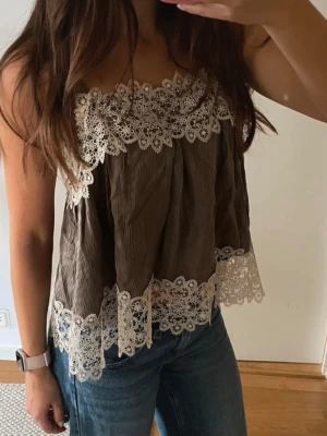  Ny populärt Brun spetsprydd linnetopp från Zara - Supersöt brun linnetopp från Zara Woman med breda spetsdetaljer i vitt längs överkant och nederkant. Toppen är ärmlös och har en luftig, loose passform som ger en chill vibe. Perfekt att styla med jeans eller kjol för en trendig look.