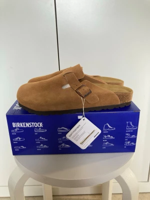 Birkenstock Boston mockatofflor brun - Snygga Birkenstock Boston tofflor i brun mocka med justerbart spänne och klassisk korksula. Perfekta för chill dagar hemma eller när du vill ha en avslappnad stil. Mjuk innersula för extra komfort och tidlös design som alltid funkar. Storlek 41