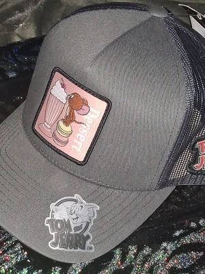 Tom & Jerry Keps  - Snygg svart keps med nät på sidorna och ett färgglatt Tom & Jerry-märke framtill. På framsidan finns ett broderat patch med Jerry och texten 'Dessert' samt milkshake och macarons. 
