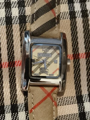 Burberry klocka med rutigt band - Burberry klocka med fyrkantig silverfärgad boett och klassiskt beige rutigt armband. Urtavlan har Burberrys logga och mönster i beige, svart och rött. Klockan är Swiss Made och har detaljer i rostfritt stål.