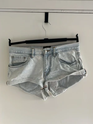 Ljusblå denimshorts med upprullad kant - Säljer ett par ljusblå denimshorts med upprullad kant och klassisk femficksdesign. Shortsen har knappgylf och bälteshällor, perfekta för varma dagar. Snygg och avslappnad passform som passar till det mesta.