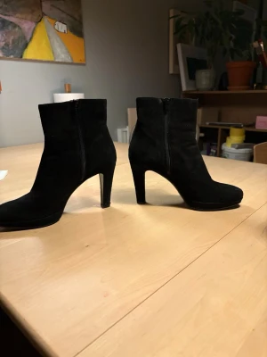Rizzo stövletter  - Snygga svarta stövletter i mocka med smal klack och dragkedja på sidan. Klassisk och stilren design som passar till många olika outfits. Perfekt höjd på klacken för att ge lite extra längd utan att bli obekvämt.