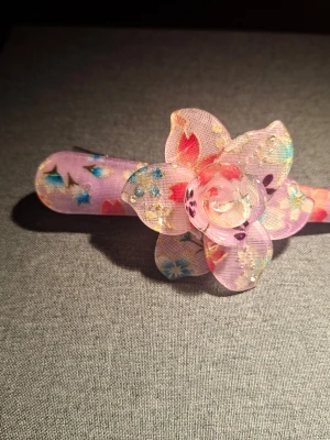 Lila hårklämma med blomma - Superfin hårklämma i lila plast med en stor dekorativ blomma i tyg. Blomman har flera lager med blommönster i rosa, blått och guldiga detaljer. Perfekt accessoar för att piffa upp frisyren med en söt och färgglad touch.