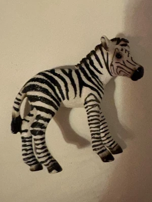 Schleich Zebra - Säljer en realistisk zebrafigur i plast med svartvita ränder och detaljerad form. Perfekt som dekoration, leksak eller samlarobjekt för dig som gillar djur och safari. Figuren har tydliga ränder och står stadigt på fyra ben.