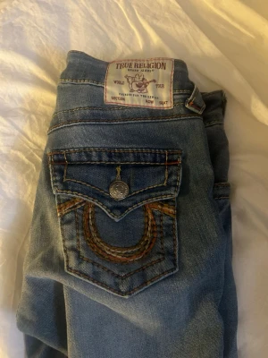 Blå True Religion jeans bootcut mid rise  - Säljer ett par blå jeans från True Religion med bootcut-modell. Jeansen har klassiska kontrastsömmar och detaljerade bakfickor med lock och knapp. Materialet är denim i bomull och passformen är rak med lätt utsvängda ben. (DESSA JEANS ÄR KLIPPTA EFTER MIN LÄNGD) jag är ca 160