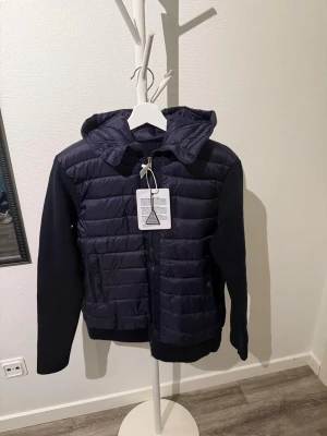 Mörkblå dunjacka med huva Moncler - Snygg mörkblå dunjacka från Moncler med quiltat mönster och stickade ärmar. Jackan har dragkedja framtill, två fickor och en praktisk huva. Perfekt för kalla vinterdagar och riktigt stilren design.