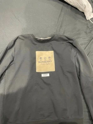 Svart sweatshirt från Burberry - Stilren svart sweatshirt från Burberry med beige patch på bröstet och tryckt logga samt texten 'London, England'. Rund halsringning och långa ärmar. Perfekt för dig som gillar klassisk streetwear med exklusiv känsla.