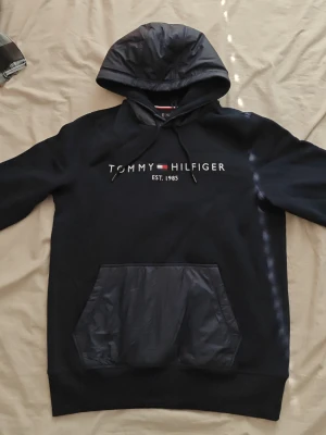 Tommy Hilfiger hoodie - Snygg svart hoodie från Tommy Hilfiger med broderad logga och text på bröstet. Huva och magficka i glansigt material ger en cool kontrast. Klassisk passform med långa ärmar och ribbade muddar. Perfekt för en avslappnad streetwear-look.