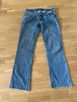 Blå bootcut jeans med rå kant - Säljer ett par blå bootcut jeans med rå, fransig kant nedtill. Jeansen har klassisk femficksmodell och en liten dekorativ dragkedja på ena fickan. Perfekta för en avslappnad och trendig look.