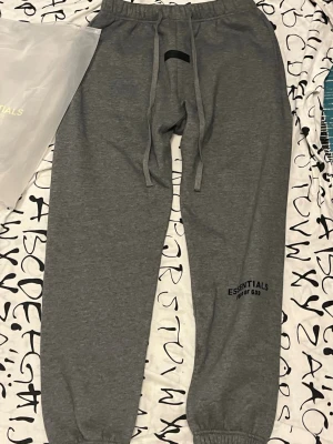 Grå Essentials mjukisbyxor - Säljer ett par grå mjukisbyxor från Essentials Fear of God. Byxorna har resår i midjan med dragsko, muddar vid bensluten och tryckt logga på benet. Perfekta för en avslappnad och trendig look. Materialet är mjukt och bekvämt, troligen bomullsblandning.