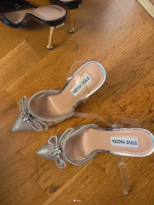 Glittriga pumps med rosett Steve Madden - Säljer ett par glittriga pumps från Steve Madden med spetsig tå, transparenta remmar och dekorativa rosetter framtill. Skorna har en klar plastklack och är täckta av små strassdetaljer som ger extra bling. Perfekta för dig som vill sticka ut!