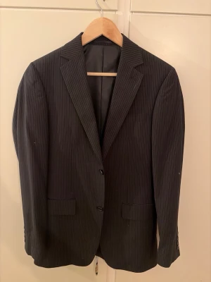 Mörkblå randig kavaj - Snygg mörkblå kavaj med tunna vita ränder. Klassisk modell med två knappar framtill, fickor och slits baktill. Perfekt för dig som vill ha en stilren och tidlös look.