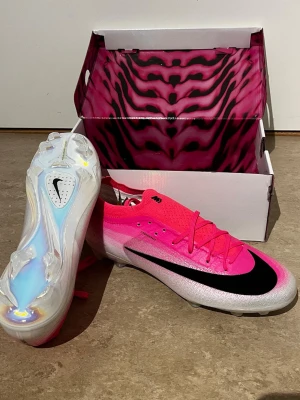 nike mercurial vapor 17 elite fg breakout - rosa/vit/svart - Säljer ett par snygga fotbollsskor från Nike i en grymt cool rosa färg med svart swoosh på sidan. Skorna har vita och skimrande detaljer samt genomskinlig sula. Perfekta för dig som vill sticka ut på planen! Skokartong med rosa mönster medföljer. Kommer utan original boxen