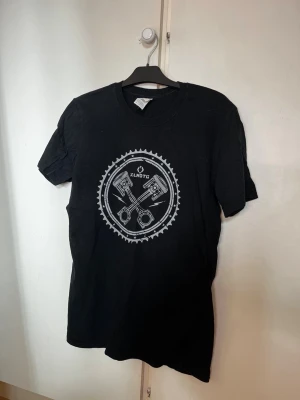 Svart t-shirt med motortryck - Svart t-shirt med ett stort tryck av två kolvar och kugghjul på bröstet. Trycket är i vitt och har texten 'KLNDTG' i mitten. Klassisk passform och rund halsringning. Perfekt för dig som gillar motorer eller vill ha en cool grafisk tee.