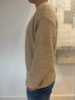 Beige stickad tröja med rund hals - En enkel och stilren beige stickad tröja med rund halsringning och långa ärmar. Tröjan har ribbade muddar vid ärmslut och nederkant. Perfekt för lager på lager och passar till det mesta. Materialet ser mjukt och bekvämt ut.