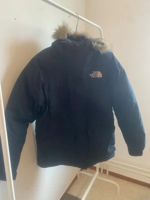 The North Face McMurdo Parka Dunjacka - Snygg svart parkas från The North Face med huva och avtagbar fuskpäls. Jackan har en klassisk längre modell, dragkedja framtill och logga på bröstet. Perfekt för kalla dagar och riktigt varm. Passar dig som vill ha både stil och funktion.
