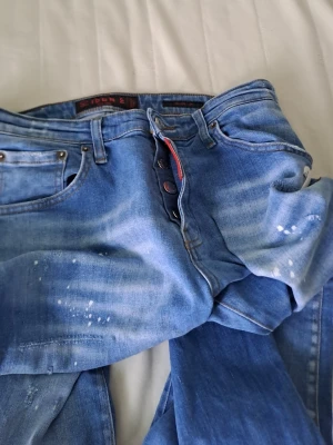 Ljusblå jeans med slitningar - Säljer ett par ljusblå jeans med slitningar och färgstänk för en cool, avslappnad look. Jeansen har raka ben, knappgylf och klassiska fem fickor. Perfekta för dig som gillar streetwear och vill ha en unik stil.