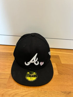 New Era Atlanta Braves keps - Säljer en svart New Era 59FIFTY keps med Atlanta Braves-logga i vitt framtill och ett litet hjärta bredvid. På sidan finns ett broderat World Series 1995-märke i gult och vitt. Klassisk rak skärm och broderade ventilationshål. Perfekt för dig som gillar sportig stil.