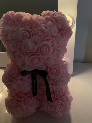 Rosa nallebjörn av rosor - Söt nallebjörn formad av ljusrosa konstgjorda rosor, dekorerad med ett svart band runt halsen. Perfekt som dekoration eller present till någon speciell. Ger ett romantiskt och gulligt intryck och passar fint i sovrum eller på skrivbord.