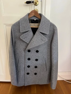 Grå dubbelknäppt rock från Burberry - Stilren grå rock från Burberry i ullmix med bred krage och dubbelknäppning framtill. Rocken har svarta knappar, två snedställda fickor och dekorativ detalj bak. Klassisk och tidlös modell som passar till många olika looks.