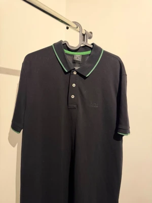 Svart pikétröja från BOSS - Svart pikétröja från BOSS med klassisk krage och gröna detaljer längs krage och ärmslut. Tröjan har tre knappar framtill och är kortärmad. Perfekt för en stilren och sportig look.