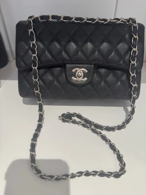 Svart handväska - Säljer en svart handväska i topp läder med silverfärgad kedja. Väskan har röd insida och klassisk rektangulär form. Perfekt för dig som vill ha en tidlös och lyxig accessoar.