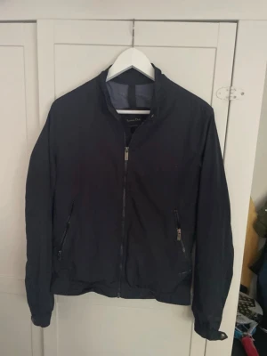 Svart vindjacka från Massimo Dutti - Stilren svart vindjacka från Massimo Dutti med dragkedja framtill och två snedställda fickor med dragkedja. Jackan har en klassisk krage och elastiska muddar vid ärmslut och nederkant. Perfekt för blåsiga dagar.