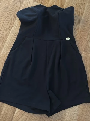 Svart axelbandslös jumpsuit - Snygg svart axelbandslös jumpsuit med korta ben och fickor på sidorna. Stilren design med markerad midja och en liten guldfärgad detalj vid ena fickan. Perfekt för dig som vill ha en enkel men trendig look.