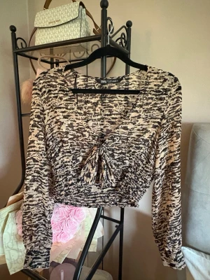 Mönstrad croppad topp från Zara - Säljer en beige och svart croppad topp från Zara med v-ringning och knyt framtill. Toppen har ett abstrakt mönster, långa ärmar och smockad midja som ger en snygg passform. Perfekt för dig som gillar trendiga detaljer och unika prints.
