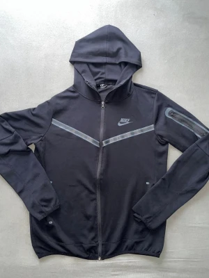 Svart Nike tech hoodie med dragkedja - Snygg svart hoodie från Nike med hel dragkedja och huva. Har gråa detaljer över bröstet och en liten ficka med dragkedja på ena ärmen. Tillverkad i mjukt och stretchigt material, perfekt för en sportig look.
