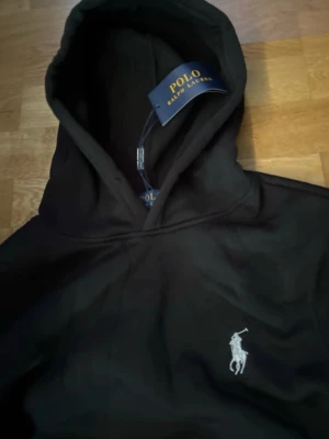 Svart hoodie från Polo Ralph Lauren - Svart hoodie från Polo Ralph Lauren med klassisk huva och broderad vit logga på bröstet. Mjuk och skön, perfekt för en avslappnad stil. Snygg och enkel att matcha med jeans eller joggers.