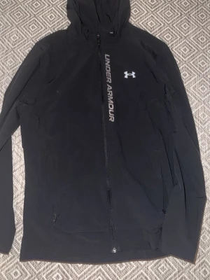 Svart vindjacka från Under Armour - Snygg svart vindjacka från Under Armour med huva och hel dragkedja framtill. Jackan har logga och text på bröstet samt två sidofickor. Tillverkad i lätt och funktionellt material som passar perfekt för aktiva dagar utomhus.