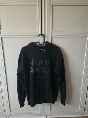 Svart hoodie med texttryck - Säljer en svart hoodie med huva och texten 'JE M'EN AMOUR' i glittrigt tryck på framsidan samt en liten stjärna. Hoodien har långa ärmar och ribbade muddar vid ärmslut och nederkant. Perfekt för en avslappnad och trendig look. Hoodien är från gina tricot💞💞