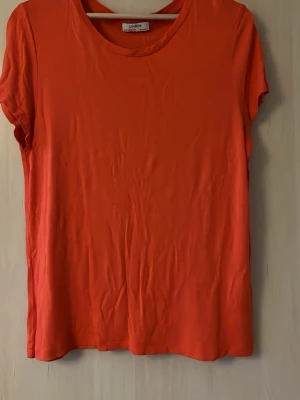 Orange t-shirt från Lindex - En enkel och snygg orange t-shirt från Lindex i mjuk bomull. T-shirten har rund halsringning och korta ärmar, perfekt till jeans eller shorts för en avslappnad look.