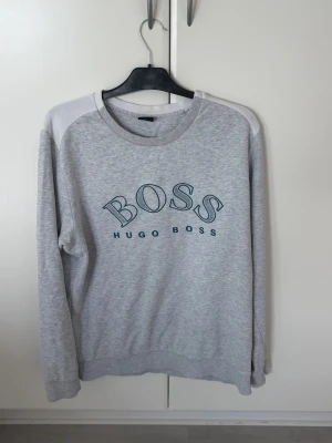 Grå sweatshirt från Hugo Boss - Snygg grå sweatshirt från Hugo Boss med stort BOSS-tryck på bröstet. Tröjan har rund halsringning, ribbade muddar och kontrastpaneler på axlarna. Perfekt för en avslappnad och stilren look. Möts upp om de är nära halmstad/göteborg