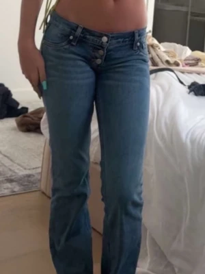 Blå bootcut jeans från Revice - Säljer ett par ljusblå bootcut jeans från Revice med låg midja och tre synliga knappar framtill. Jeansen har klassisk femficksdesign och är tillverkade i denim med lätt stretch. Perfekta för en trendig och avslappnad look.