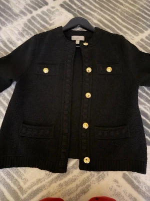 Svart cardigan med guldknappar - Stilren svart cardigan med dekorativa guldknappar framtill och fyra fickor. Klassisk rak passform och rund halsringning. Perfekt att slänga över en topp för en chic look. Använd 1 gång. Storlek xs men passar även S