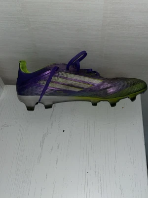 Lila och gröna fotbollsskor med dobbar - Säljer ett par snygga fotbollsskor i lila och grönt med vita detaljer. Skorna har låg profil, snörning och fasta dobbar för spel på gräs. Ovandelen är tillverkad i syntetmaterial som ger bra bollkontroll och lätt känsla på foten. Perfekta för dig som vill sticka ut på planen.