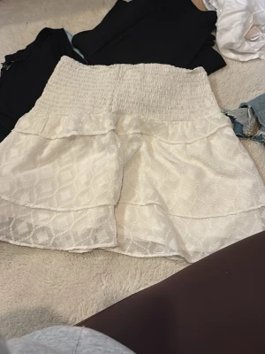 Vit volangkjol med smock - Söt vit kjol med smock i midjan och dubbla volanger. Kjolen har ett diskret mönster i tyget och är gjord i ett lätt, luftigt material som passar perfekt till sommaren. Den har en kort, A-linjeformad siluett.