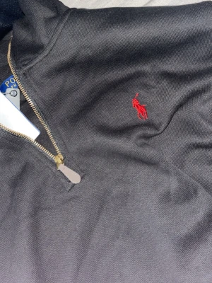 Polo ralph lauren Quarter zip - Svart långärmad tröja med half-zip och hög krage från Polo Ralph Lauren. Tröjan har det klassiska röda broderade logot på bröstet och är tillverkad i ett mjukt material. Perfekt för en stilren och avslappnad look.