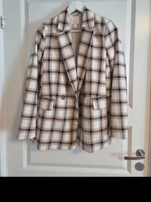 Snygg rutig kavaj från H&M – Perfekt skick! - En jättefin, dubbelknäppt kavaj i ett klassiskt rutmönster med bas i beige/off-white och linjer i svart och brunt. Otroligt mångsidig modell som passar perfekt till både vardags och fest! Rak och modern passform, dubbelknäppt med snygga knappar fram och fickor med lock.       Superfin att matcha både till jeans eller kjol.