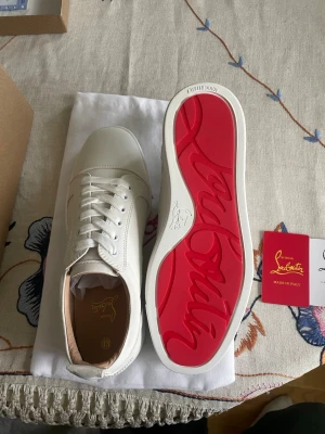 Vita sneakers från Christian Louboutin - Stilrena vita sneakers från Christian Louboutin med klassisk röd sula och diskret logga på sidan. Skorna har vita skosnören, rund tå och är tillverkade i slätt läder. Perfekta för dig som vill ha en lyxig och trendig look.
