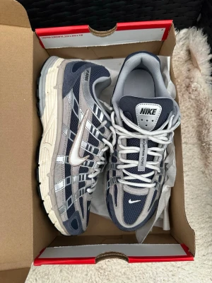 Nike grå/blå sneakers Air Max - Nike sneakers i grått och marinblått med vita detaljer och meshpaneler. Klassisk swoosh-logga på sidan och retroinspirerad design. Skorna har vita snören och en robust sula i beige och svart. Perfekta för dig som gillar sportig och trendig stil.