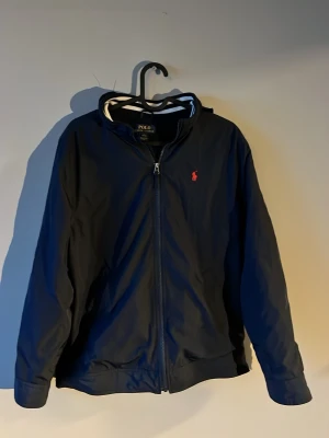 Mörkblå vindjacka från Polo Ralph Lauren - Snygg mörkblå vindjacka med huva från Polo Ralph Lauren. Jackan har dragkedja framtill, ribbade muddar och en röd logotyp på bröstet. Perfekt för blåsiga dagar och enkel att matcha med olika outfits. Stolek 14-15 år L i barn modellerna från Ralph lauren 