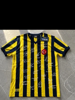 Fenerbahçe gul och blå matchtröja - Säljer en Fenerbahçe matchtröja i gult och mörkblått med vertikala ränder. Tröjan har V-ringning, tryckt turkisk flagga och flera autografer på framsidan. Märkt med 'TRİBÜN' och 'Fenerbahçe' på bröstet. Perfekt för fotbollsfans!