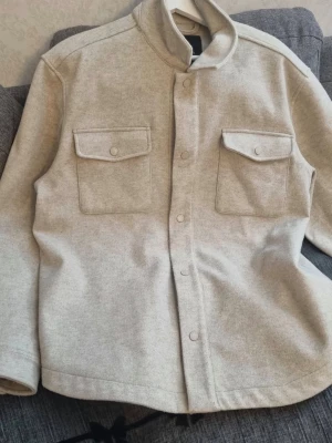 Beige jacket  med fickor - Stilren beige jacket  med två stora bröstfickor med lock och tryckknappar framtill. Rak passform och hög krage ger en modern look. Materialet ser ut att vara mjukt och lite tjockare, perfekt för lager på lager. Enkel att matcha till olika outfits.